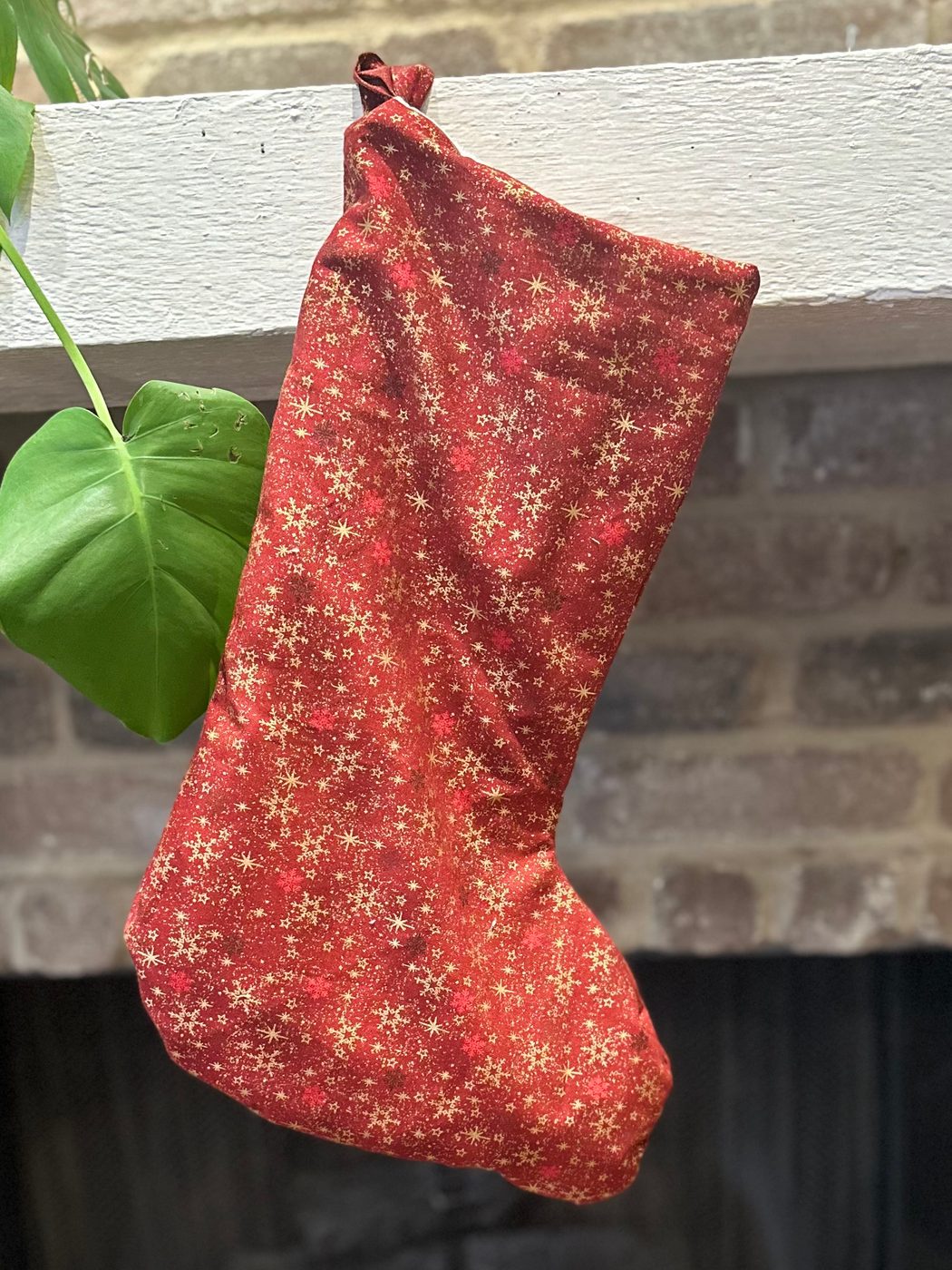 A handmade Christmas stocking in deep red cotton scattered with gold snowflakes and stars, hanging from a white mantel above a brick fireplace