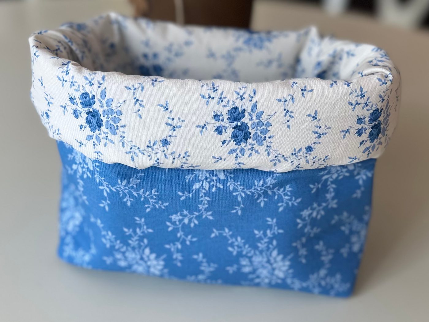 A handmade fabric storage basket in cornflower blue florals, its cuff folded down to show a white and blue rose lining